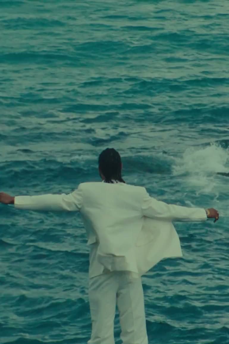 Man in a white suit wading into the sea with his arms spread