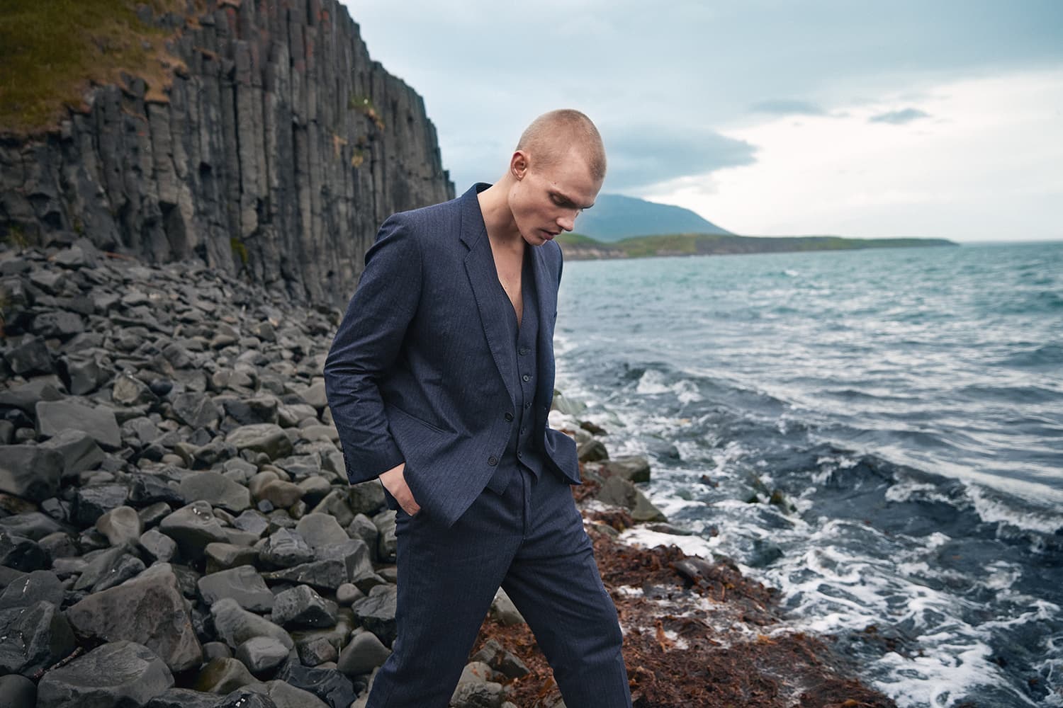 Man in a navy suit on a rocky shore under basalt cliffs